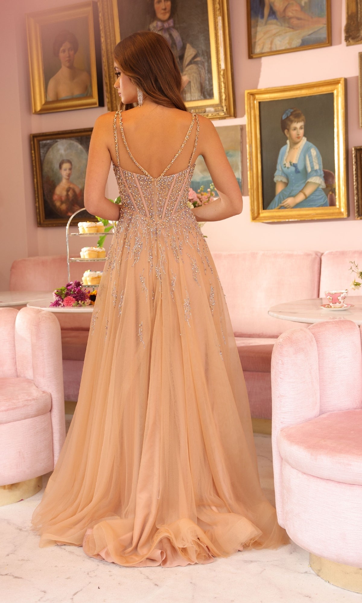 A woman in the Ava Presley 40113 Long A-Line Prom Dress, featuring a beaded corset bodice, stands with her back to the camera in a pink vintage-style room decorated with framed portraits.